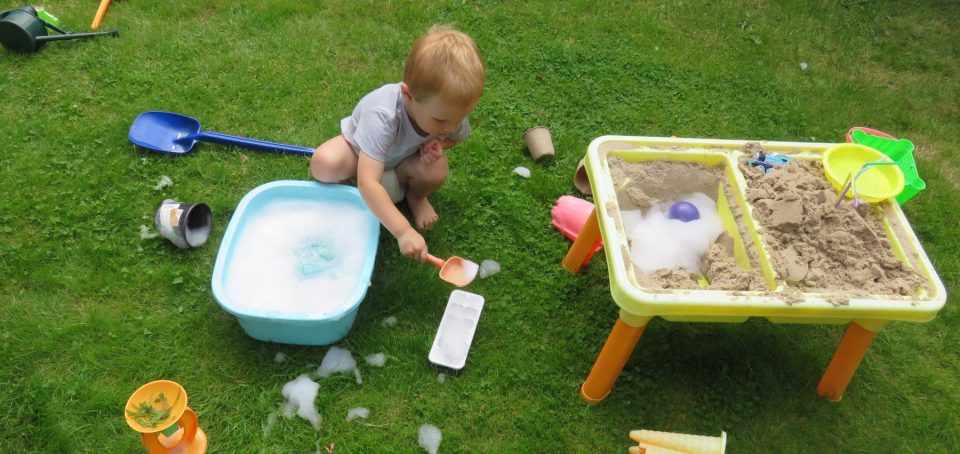 child on lawn with a sand and water table plus lots of toys and foamy water in a bowl