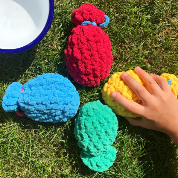 four reusable water balloons on the grass with a child's hand grabbing one. A bowl of water in the top left corner