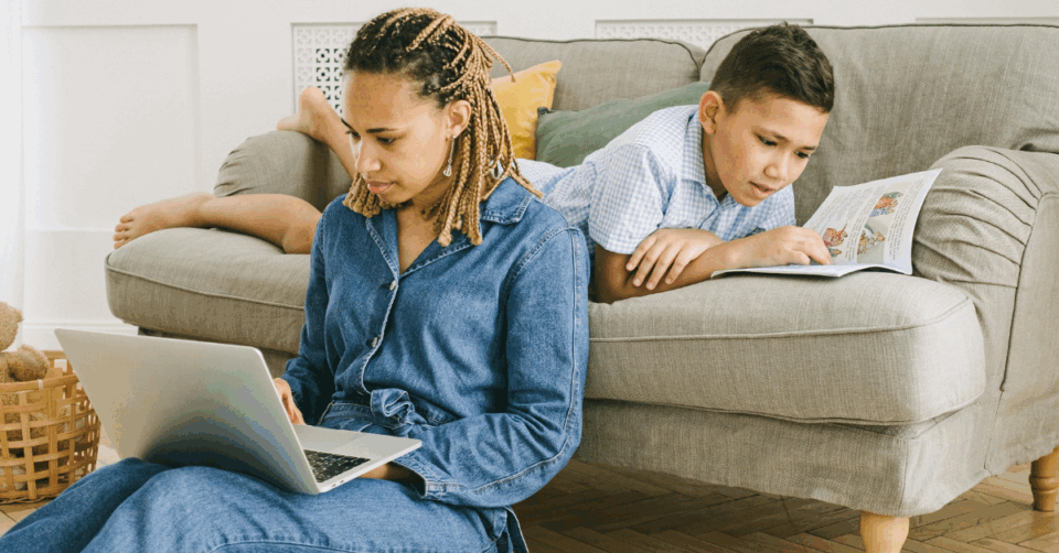 a mum working on a laptop whilst child lays on sofa behind her