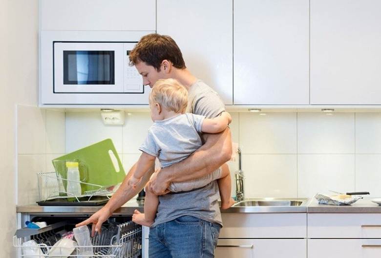 a dad holding a baby whilst loading a dishwasher