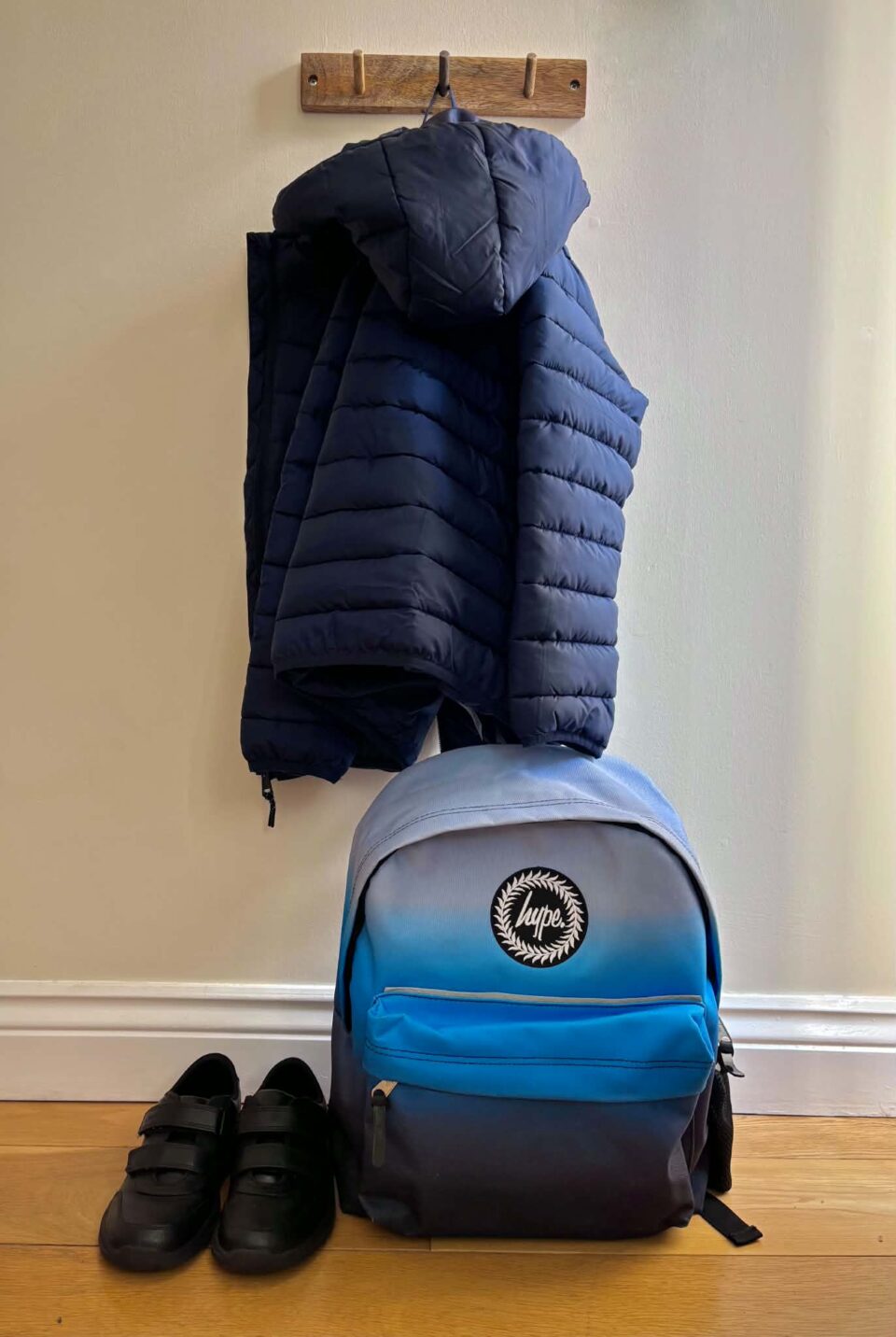 a kids winter coat hanging on a hook with a school bag below next to black boys school shoes