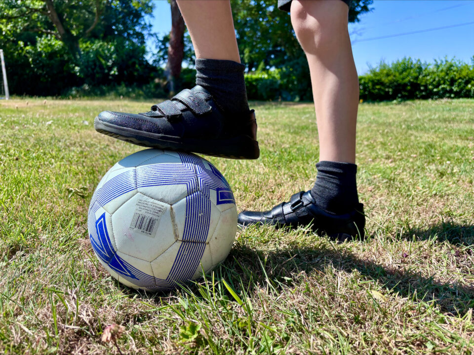 kid wearing black velcro school shoes with one foot up on a football