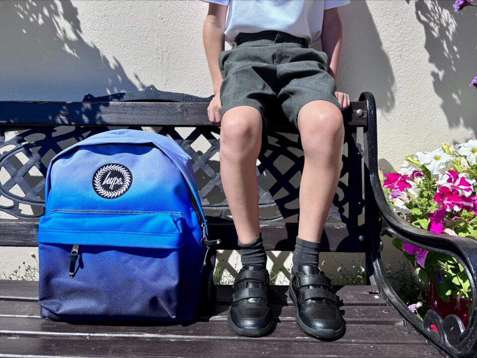 a child sat on a bench with a backpack, school shoes and shorts plus a white top on